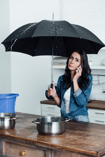 Sad woman talking on smartphone and holding umbrella under leaking ceiling in kitchen - Photo, Image