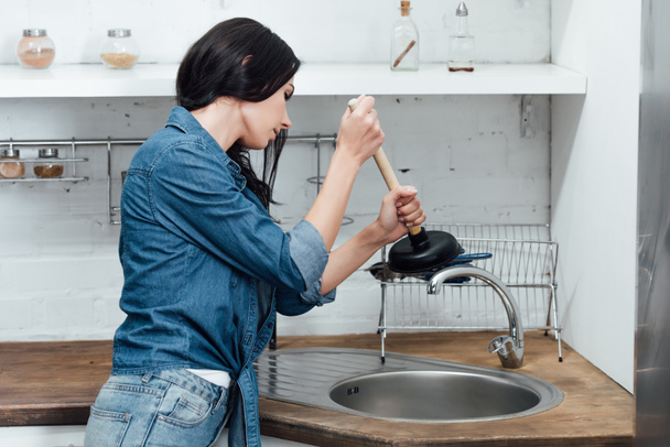 Concentrated woman in denim shirt using plunger in kitchen - Fotó, kép