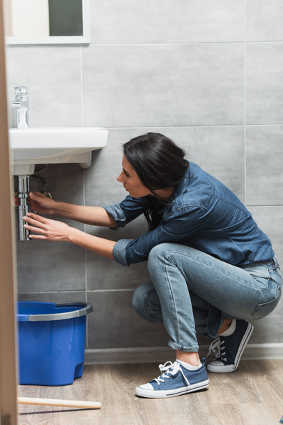Brunette woman in jeans fixing pipe in bathroom - Foto, imagen
