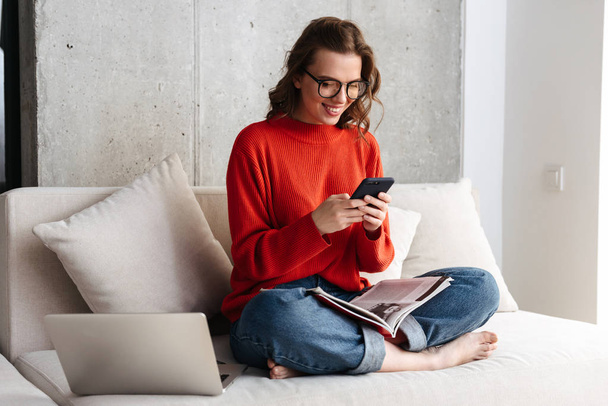 Cheerful young casually dressed woman in headphones sitting on a couch at home, studying with laptop computer, holding mobile phone - 写真・画像
