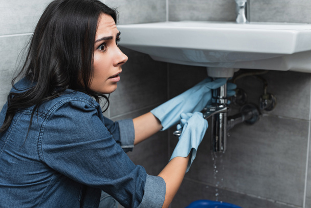 Preoccupata ragazza bruna in camicia di jeans riparazione tubo lavandino con chiave
 - Foto, immagini