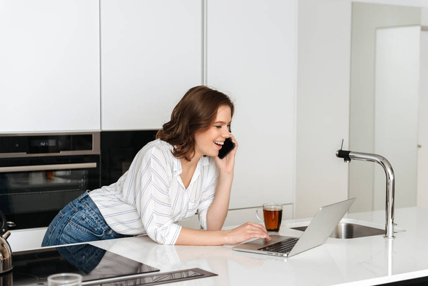 Szczęśliwy młody kobieta stoi w kuchni, w domu, przy użyciu telefonu komórkowego - Zdjęcie, obraz