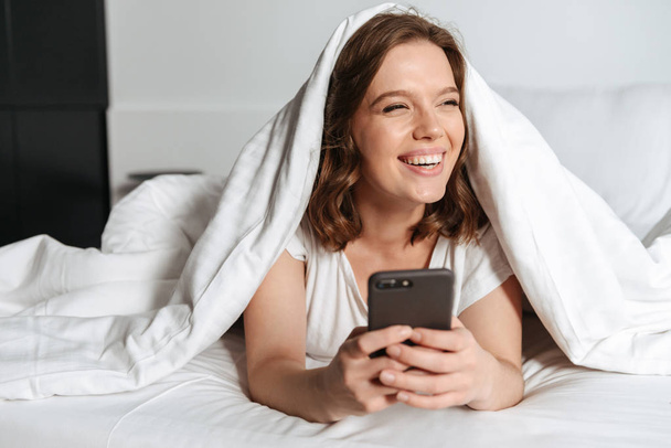 Attractive smiling young woman lying in bed under the blanket at home, using mobile phone - Фото, зображення