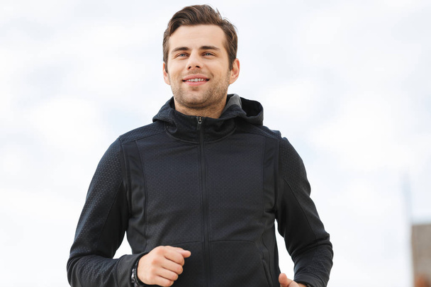 Image of happy sporty man 30s in black sportswear running along pier at seaside - Photo, Image