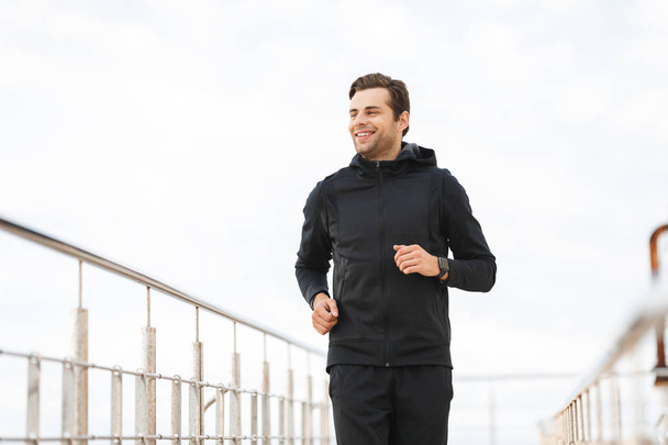Image of joyful sporty man 30s in black sportswear running along pier at seaside - Φωτογραφία, εικόνα