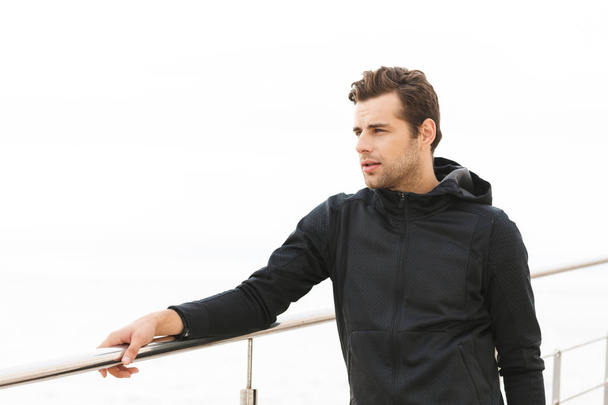 Image of attractive sporty man 30s in black sportswear standing on pier at seaside - Photo, image