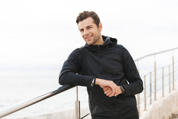 Image of happy sportsman 30s in black tracksuit standing on pier at seaside - Photo, image