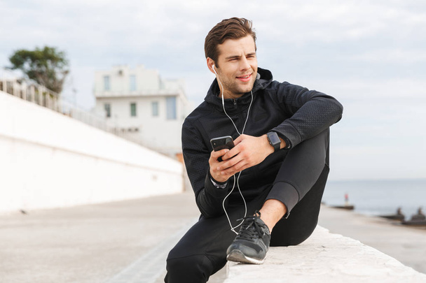 Image of european sportsman 30s in black sportswear using earphones and mobile phone while sitting on boardwalk at seaside - Foto, immagini