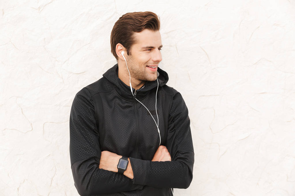 Image of attractive sportsman 30s in black sportswear and earphones standing over white wall outdoor - Foto, Imagen