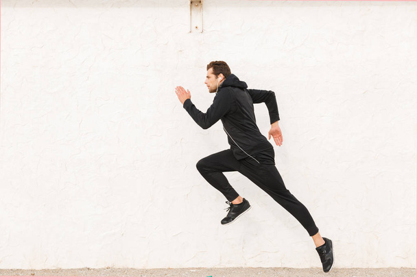 Image of sportive man 30s in black sportswear and earphones running along white wall outdoor - Fotó, kép