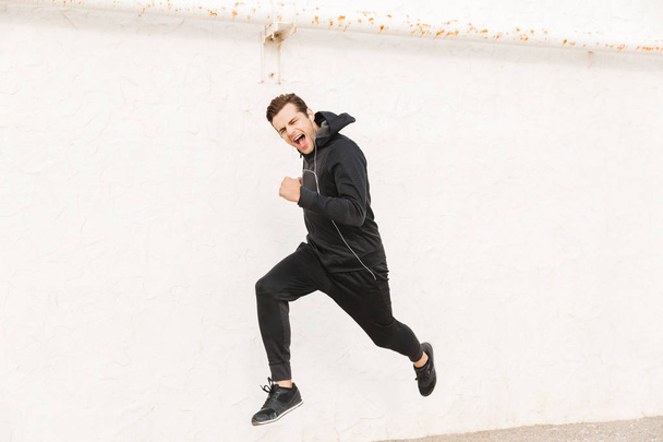 Image of joyful man 30s in black sportswear and earphones running along white wall outdoor - Fotografie, Obrázek