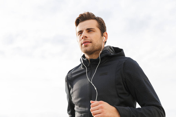 Image of athletic guy 30s in black sportswear and earphones working out and running by seaside - Фото, зображення