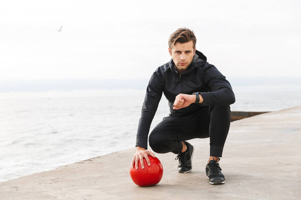 Handsome confident young sportsman working out at the beach, doing exercises with a heavy ball, looking at smartwatch - Foto, afbeelding