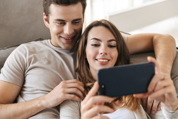 Happy young couple sitting on a couch at home, using mobile phone - Фото, зображення
