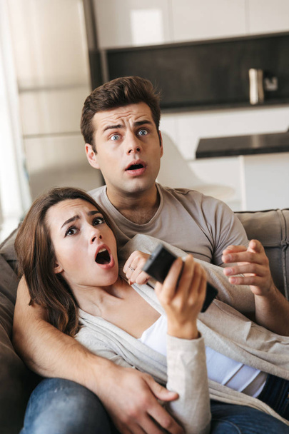 Scared young couple sitting on a couch at home, watching TV - Foto, Imagen