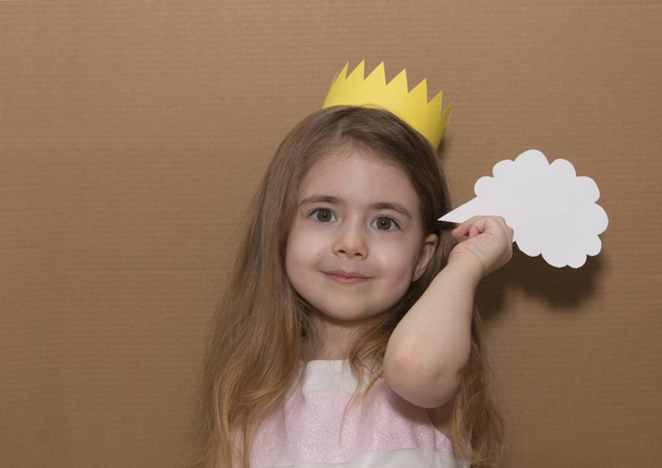 Close up portrait little girl with a crown standing isolated over cardboard background holding speech bubble. Handmade. space for text - Photo, Image
