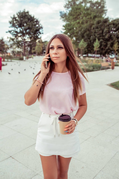 A girl stands in the summer in city. In his hand, the smartphone calls on the phone with a cup of tea or coffee. Woman in casual clothes. - Foto, Bild
