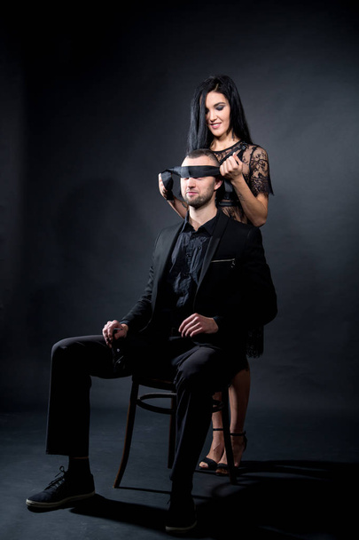 Lovers role-playing games. Girl dressed in black lacy negligee, wearing sexy underwear. Dominate obey undress seduce a partner, boss seating on chair blindfold. Sensual date idea. Thematic bdsm party - Fotoğraf, Görsel
