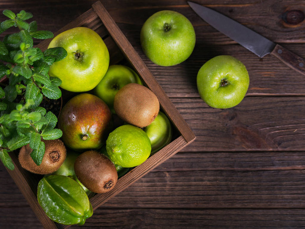 Healthy food background. Concept of healthy food. Fresh green fruits in box on dark wooden background. Set of green fruits for healthy diet and detox: apple, lime, kiwi, carambola and mint. Flat lay - Photo, Image