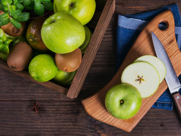 Concept of healthy food. Fresh green fruits in a box, green apple on cutting board. Dark wooden background. Set of green fruits for healthy diet and detox: apple, lime, kiwi, mango, carambola and mint - Photo, Image