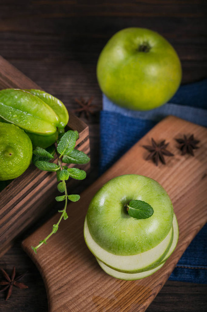 Concept of healthy food. Sliced green apple on cutting board and fresh green fruits in a box. Dark wooden background. Set of green fruits for healthy diet and detox: apple, lime, carambola and mint - Fotó, kép
