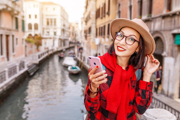 Young happy asian traveler woman with smartphone on the pier of a narrow canal and street in Venice. Vacation in Italy concept - Fotó, kép