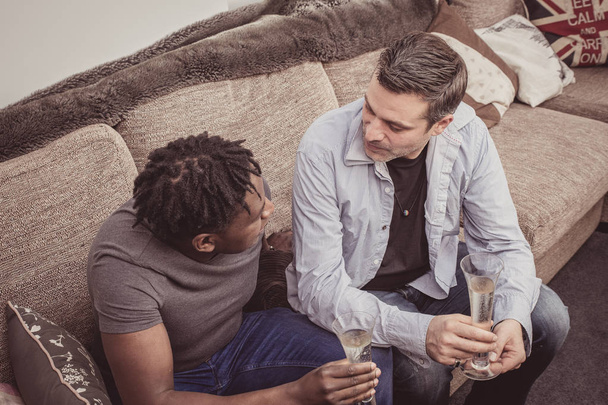 Interracial gay couple enjoy some champagne at home on the sofa.  - Фото, зображення
