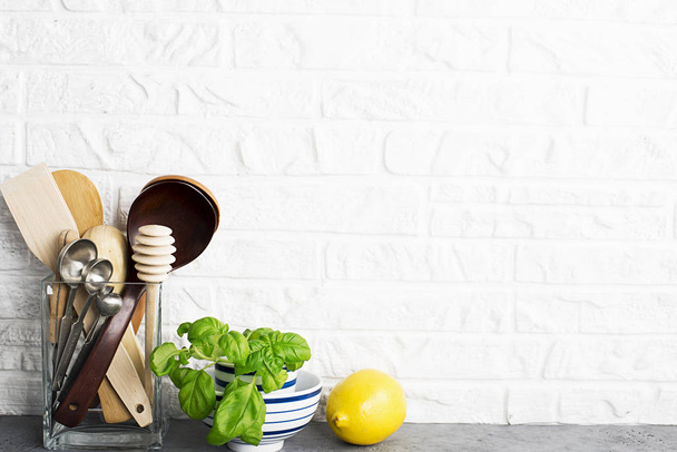 Kitchen tools, olive cutting board on a kitchen shelf against a white brick wall. Selective focus. Fresh basil and lemon. White and black modern cutlery. No plastic house - Zdjęcie, obraz