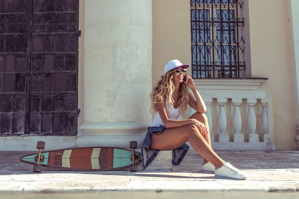Beautiful girl in the summer city. Sits near building in a row of skateboard. Phone calls happy laughs with long hair. Free space for text. - Fotó, kép