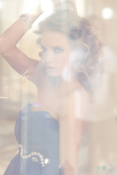 Portrait of a charming woman with stylish curly hairstyle wearing a blue dress posing next to a mirror. Beauty, vogue, fashion. - Photo, Image