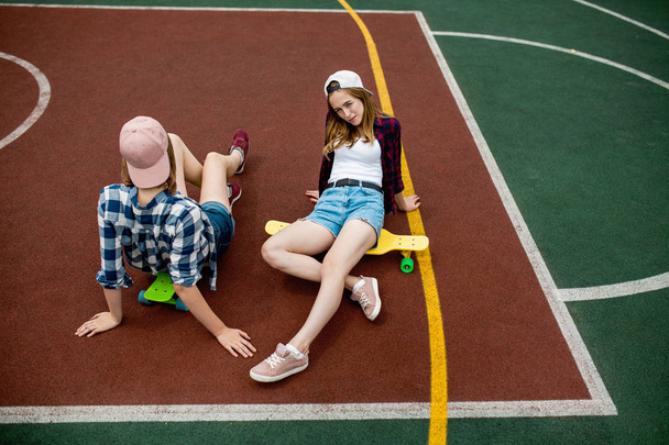 Two pretty blond girls wearing checkered shirts, caps and denim shorts are sitting on the bright longboards on the sportsfield. Beautiful legs in a good fit. Sport and cool style. - Φωτογραφία, εικόνα