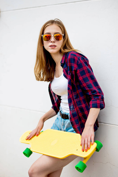 A pretty blond girl wearing sunglasses, checkered shirt and denim shorts is standing in front of the gray wall and holding a yellow longboard in front of her. Cool style. - Foto, Imagen