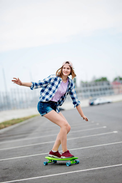A beautiful smiling blond girl wearing checkered shirt, cap and denim shorts is longboarding while stretching out her hands. Sport and cool style - Foto, Imagen