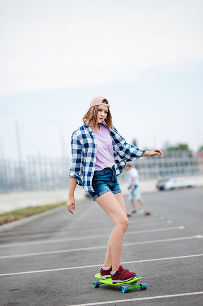 A beautiful blond girl wearing checkered shirt, cap and denim shorts is longboarding while stretching out her hands. Sport and cool style - Fotoğraf, Görsel