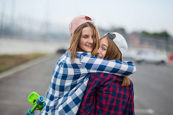 Two pretty smiling blond girls wearing checkered shirts, caps and denim shorts are standing and hugging on the empty car park with longboards in their hands. Sport and cool style - Zdjęcie, obraz