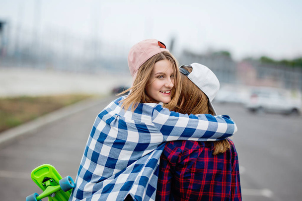 Two pretty smiling blond girls wearing checkered shirts, caps and denim shorts are standing and hugging on the empty car park with longboards in their hands. Sport and cool style. - Φωτογραφία, εικόνα