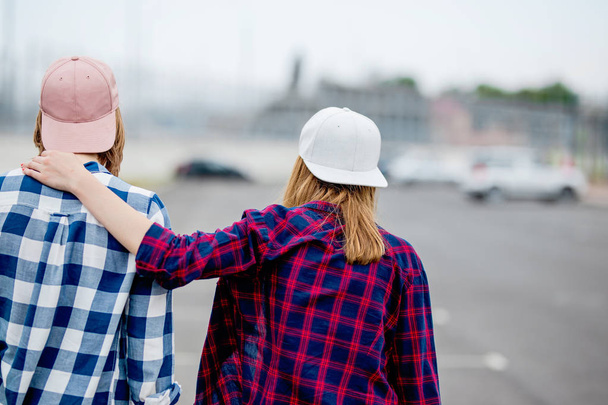 Two blond girls wearing checkered shirts, caps and denim shorts are standing with their backs on the empty car park. Sport and cool style. - Foto, Imagem