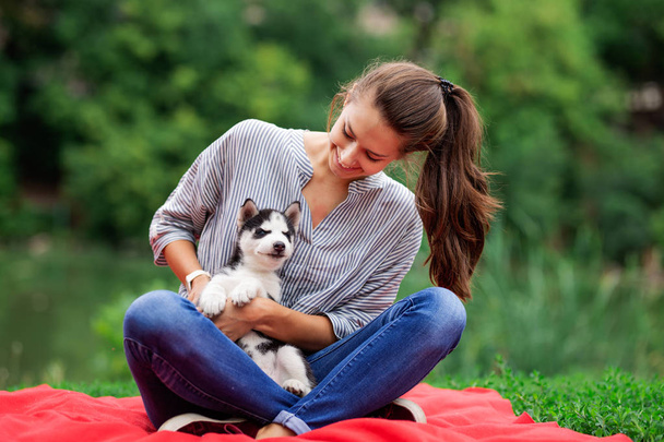 A beautiful smiling woman with a ponytail and wearing a striped shirt is playing with a sweet husky puppy while resting on the red blanket on the lawn. Love and care for pets. - Foto, imagen