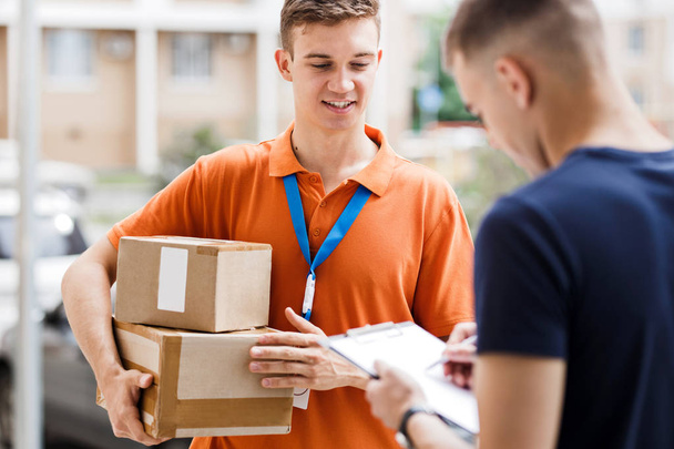 A smiling person wearing an orange T-shirt and a name tag is delivering a parcel to a client, who is putting his signature on the receipt. Friendly worker, high quality delivery service - Foto, imagen