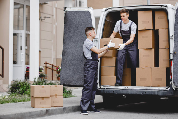 Two young handsome movers wearing uniforms are unloading the van full of boxes. House move, mover service - Photo, image