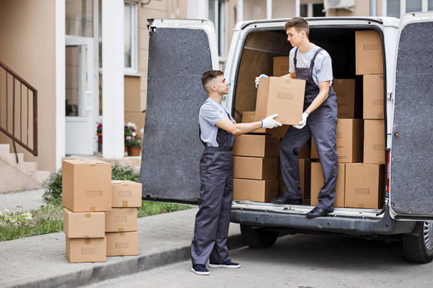 Two young handsome movers wearing uniforms are unloading the van full of boxes. House move, mover service - Fotografie, Obrázek