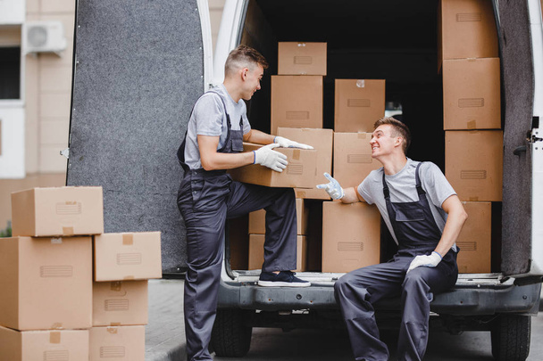Two young handsome movers wearing uniforms are laughing while unloading the van full of boxes. House move, mover service - Foto, Imagem