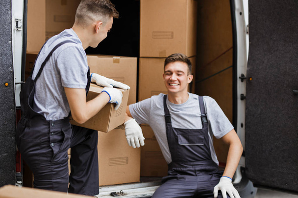 Two young handsome movers wearing uniforms are laughing while unloading the van full of boxes. House move, mover service - Foto, Bild