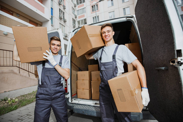 Two young handsome smiling workers wearing uniforms are standing in front of the van full of boxes holding boxes in their hands. The block of flats is in the background. House move, mover service - Foto, Imagem