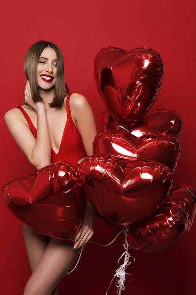 Young beautiful brunette woman with a sweet white smile in red lingerie and a balloon in the form of heart in his hands on a  red background - Zdjęcie, obraz