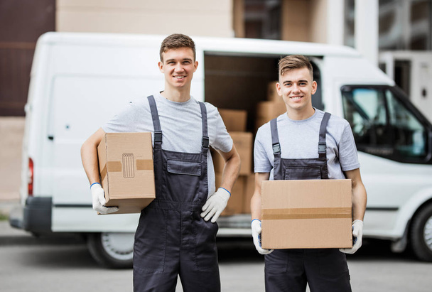 Two young handsome smiling workers wearing uniforms are standing in front of the van full of boxes holding boxes in their hands. House move, mover service - Foto, Imagen