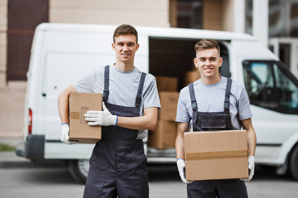 Two young handsome smiling workers wearing uniforms are standing in front of the van full of boxes holding boxes in their hands. House move, mover service - Foto, imagen
