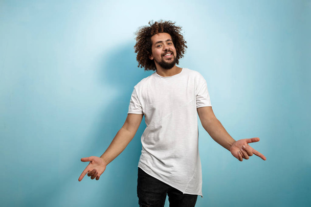 A curly-headed brunet man is stretching out his arms with point fingers expressing enjoyment. Asymmetric white T-shirt over the blue background. - Foto, immagini