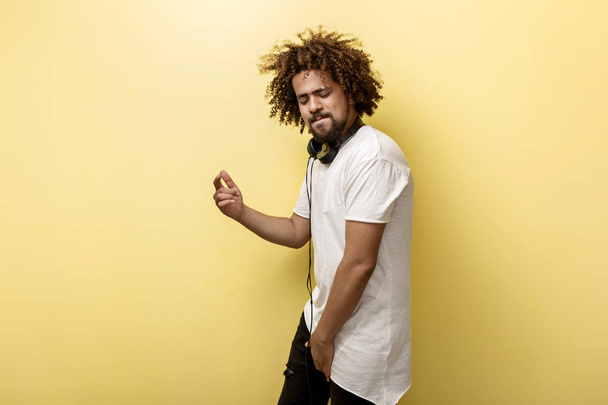 A man with his hair in a frizz and headphones on the neck is dancing with a contended smile and flying hair over the yellow background - Photo, Image