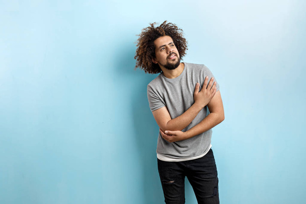 A curly-headed handsome man wearing a gray T-shirt and ripped jeans is standing and thinking with his hand on the chest, looking upwards over the blue background - Foto, Imagem